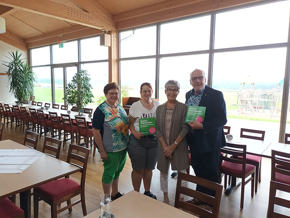 Foto von drei Frauen und einem Mann in einer Gaststätte. Der Mann und eine Frau halten eine grüne 7 %-Broschüre in den Händen.