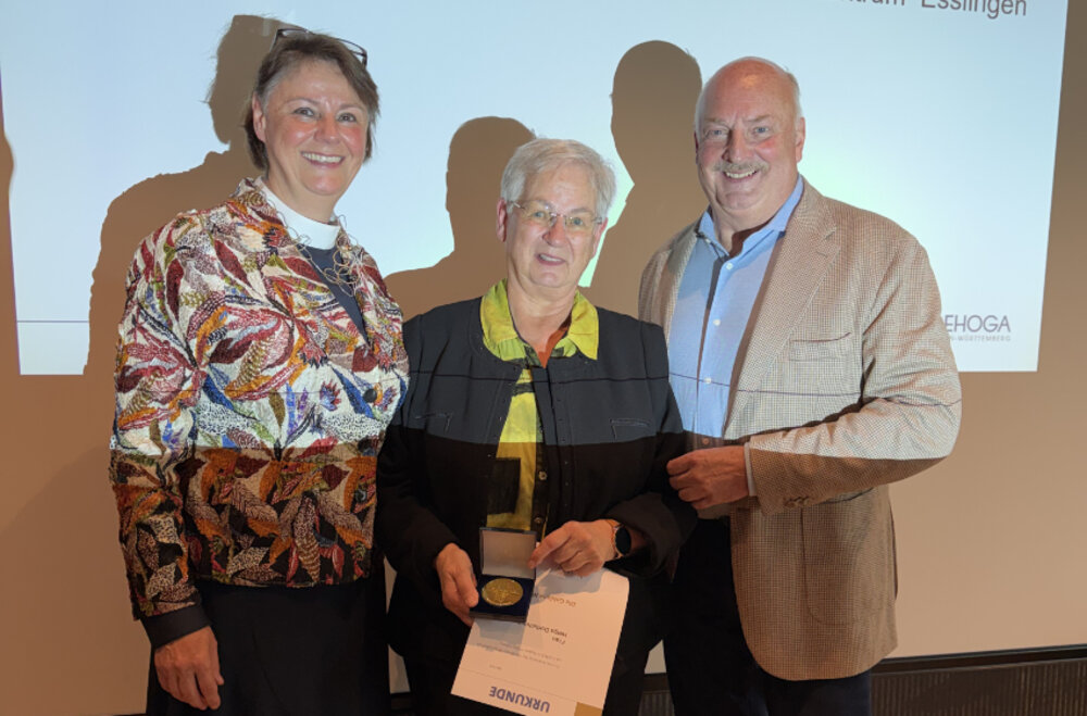 Zwei Frauen und ein Mann lächeln in die Kamera. Sie stehen vor einer Leinwand. Die Frau in der Mitte hält eine Urkunde und eine Medaille in der Hand.