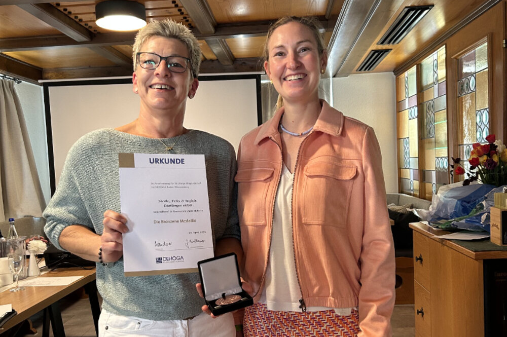Zwei Frauen posieren für ein Foto. Die Frau links hält eine Urkunde und eine Medaille in der Hand.