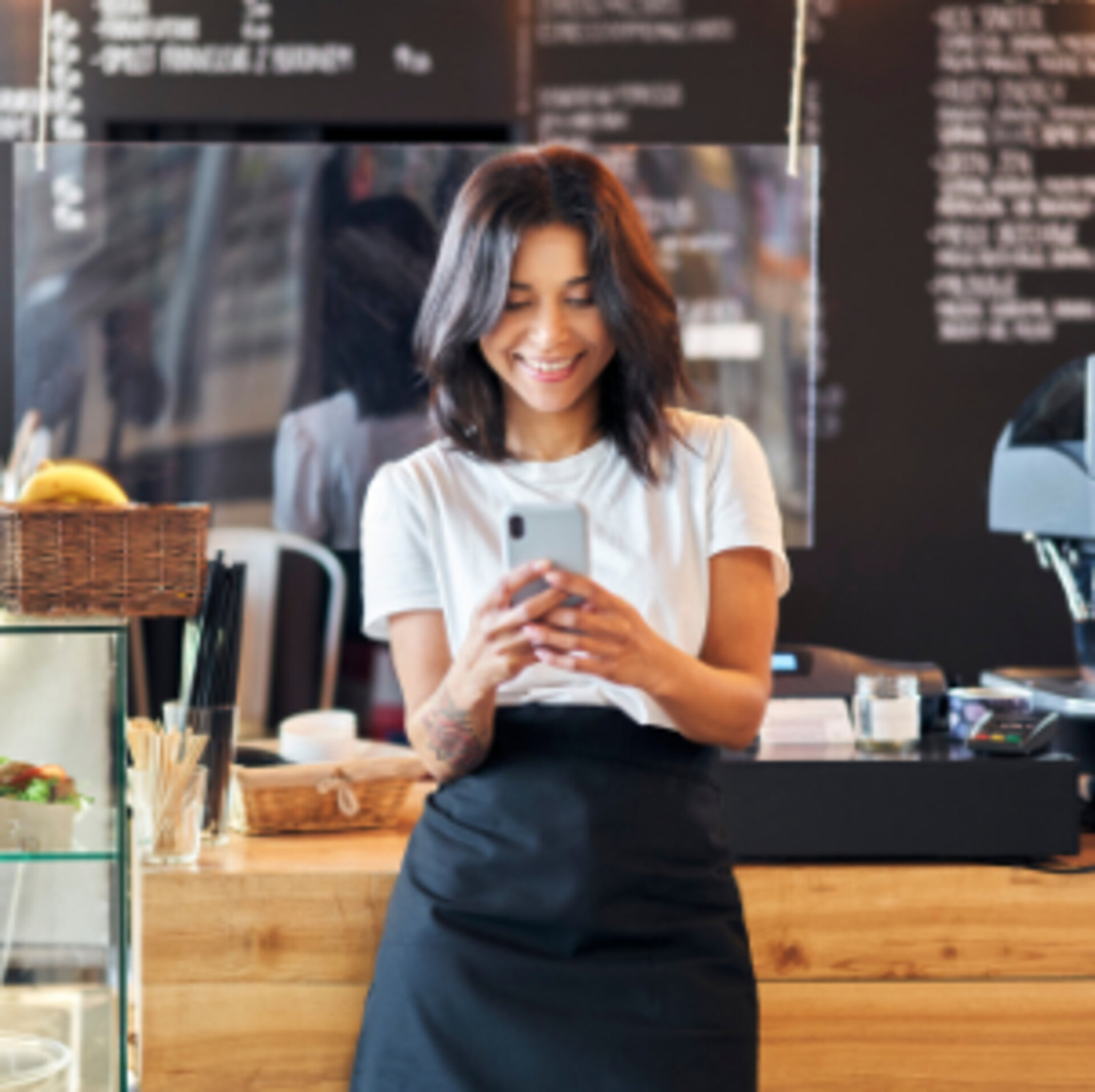 Eine Frau steht vor einer Theke in einem Café. Sie schaut lächelnd auf ihr Smartphone