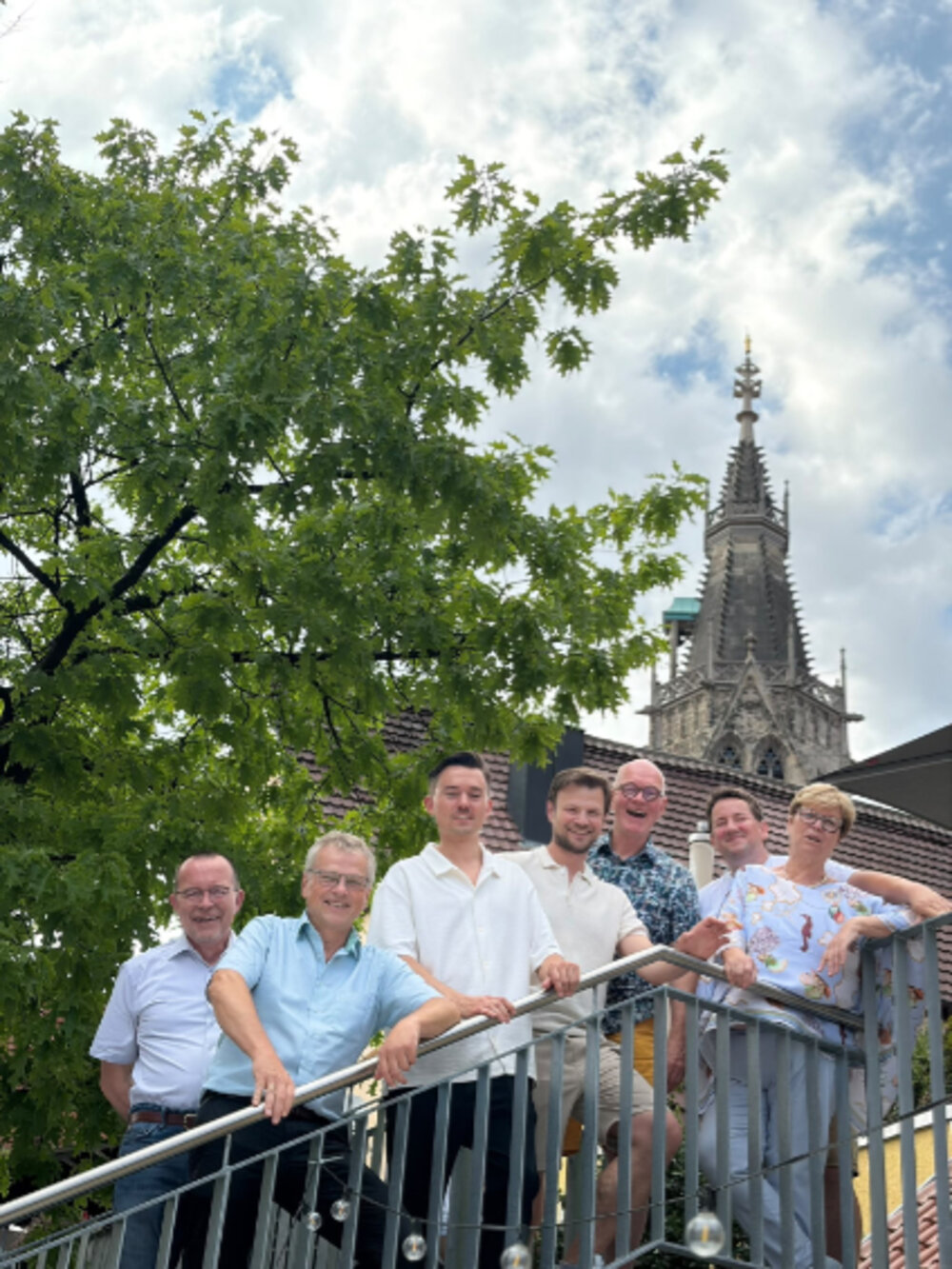 Sieben Personen lassen sich auf eine Treppe fotografieren. Im Hintergrund sieht man einen Baum und einen Kirchturm
