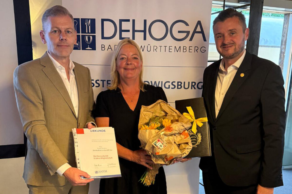 Zwei Männer und eine Frau posieren für ein Foto. Die Frau in der Mitte hält einen Blumenstrauß in den Händen. Der Mann links hält eine Urkunde