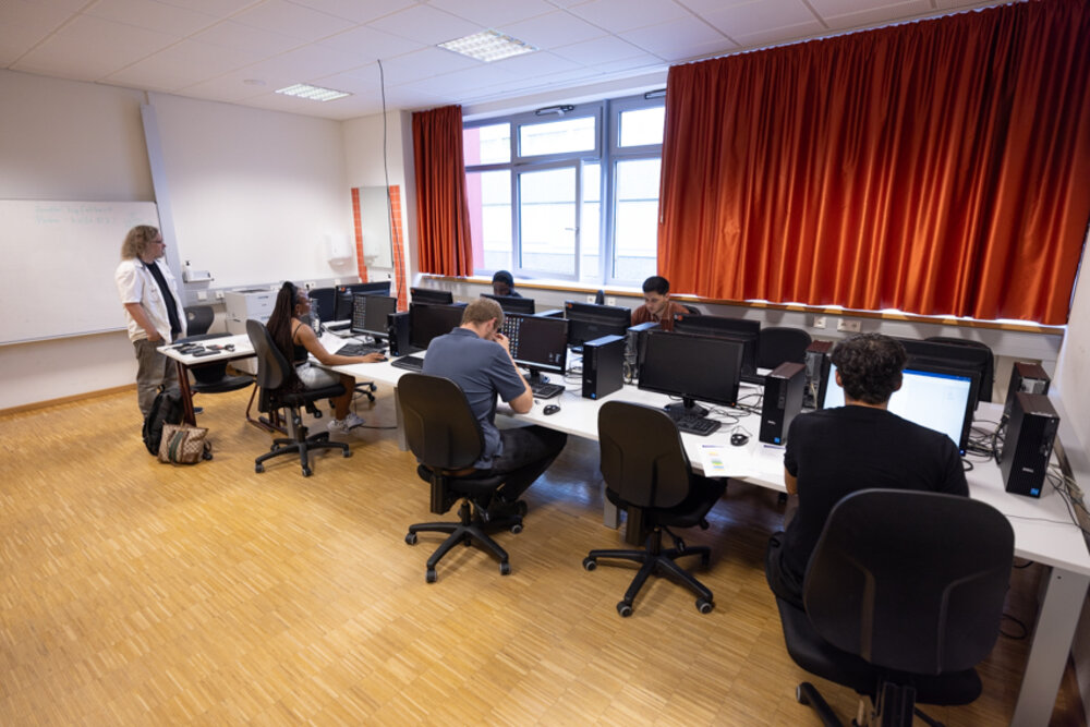 Prüfungssituation: Mehrere junge Menschen sitzen in einer Art Klassenzimmer an Tischen, vor ihnen stehen Computer. Eine Lehrerin steht am Rande des Raums