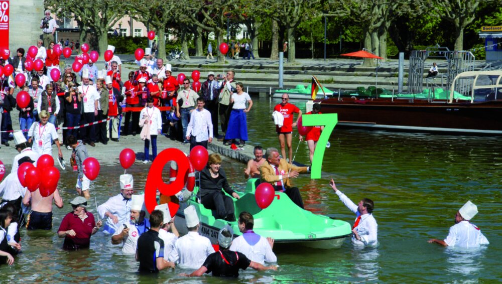 Foto von mehreren voll bekleideten Menschen, die rote Luftballons in der Hand halten und in einem See stehen.
