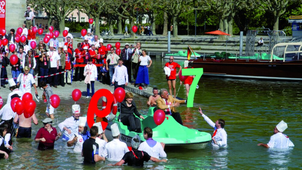 Foto von mehreren voll bekleideten Menschen, die rote Luftballons in der Hand halten und in einem See stehen.