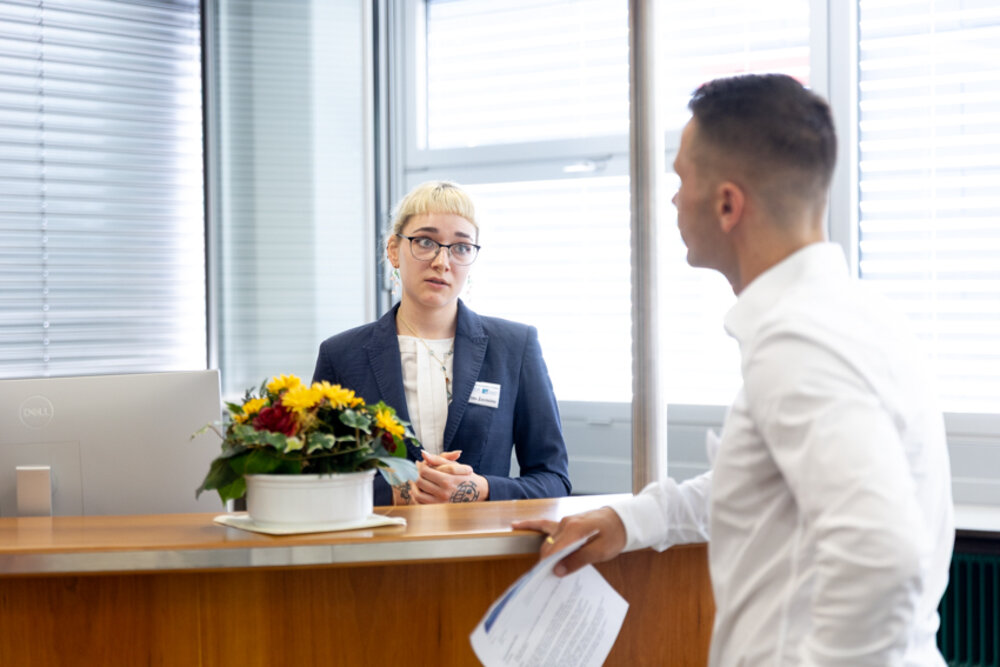 Eine junge Frau in Berufskleidung steht an einer Rezeption und spricht mit einem Gast
