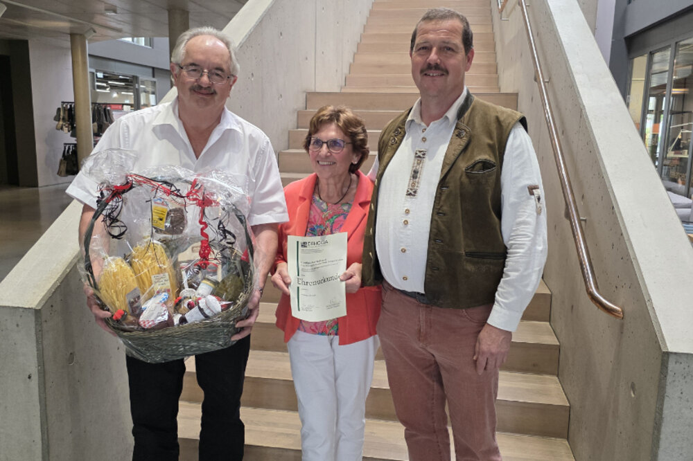 Zwei Männer und eine Frau posieren vor einer Treppe für ein Foto. Der Mann links hält einen Geschenkkorb in den Händen, die Frau in der Mitte hält eine Urkunde. Alle lächeln