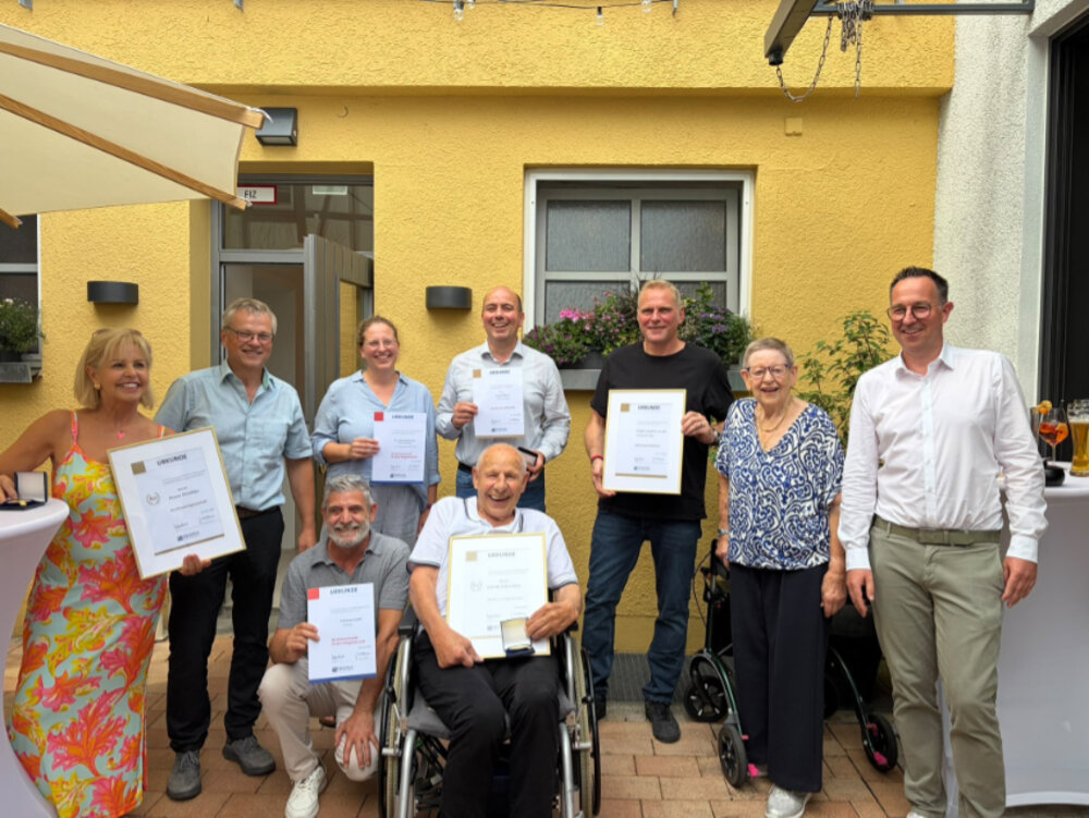 Neun Personen posieren vor einem Haus für ein Foto. Einige halten Urkunden in den Händen, alle lächeln