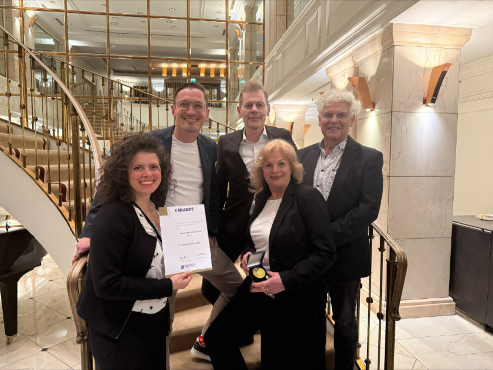 Drei Männer und zwei Frauen posieren an einer Treppe für ein Foto. Eine Frau im Vordergrund hält eine Urkunde in der Hand, die andere eine Medaille.