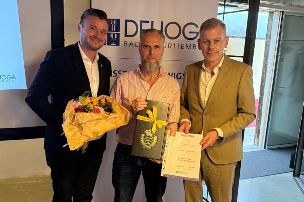 Drei Männer posieren für ein Foto. Der Mann links hält einen Blumenstrauß in den Händen. Der Mann rechts hält eine Urkunde. Der Mann in der Mitte hält ein Geschenkpaket in den Händen