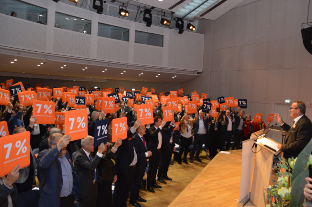 Foto von vielen Menschen in einem Saal, die Sitzkissen mit dem Druck 7 % in die Höhe halten.