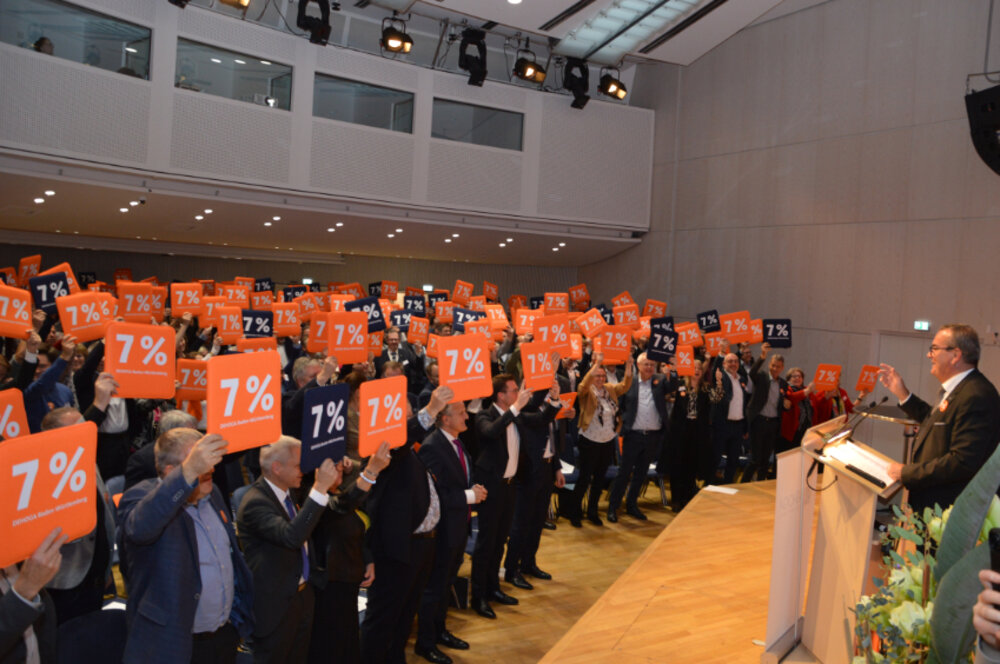 Foto von vielen Menschen in einem Saal, die Sitzkissen mit dem Druck 7 % in die Höhe halten.