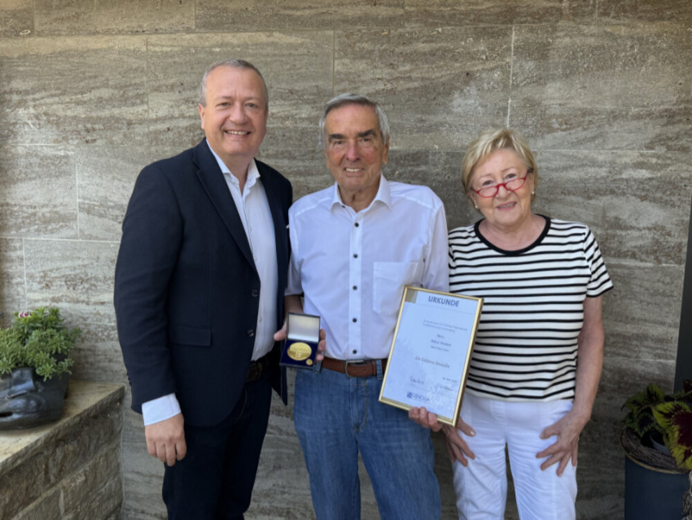 Zwei Männer und eine Frau posieren lächelnd für ein Gruppenfoto. Der Mann in der Mitte hält eine goldene Medaille und eine Urkunde in der Hand