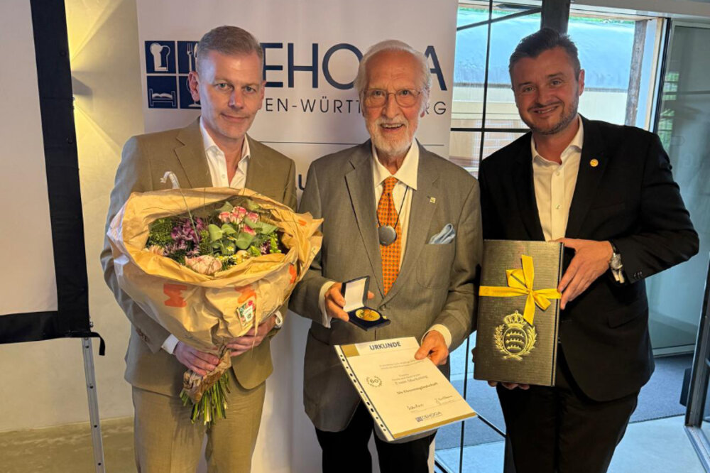 Drei Männer posieren für ein Foto. Der Mann in der Mitte hält eine Urkunde und eine Medaille in den Händen. Der Mann rechts hält ein Geschenkpaket in den Händen