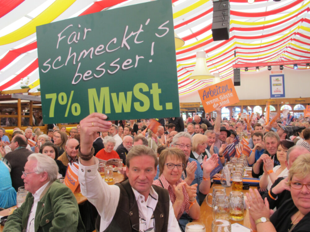 Foto eines großen Festzelts mit vielen Menschen. Im Vordergrund hält ein Mann ein Plakat mit der Aufschrift "Fair schmeckt's besser-7 %".
