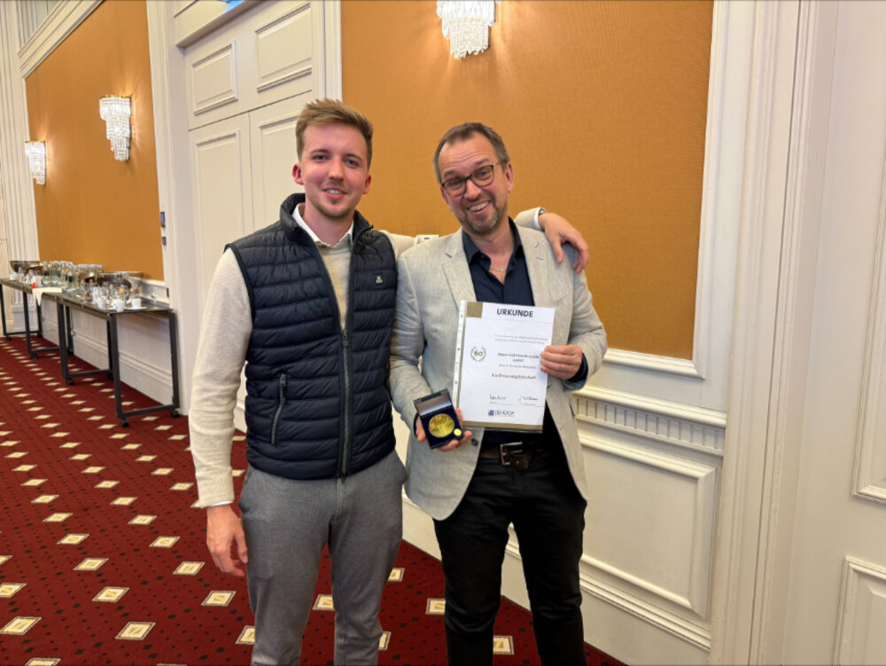 Zwei Männer lächeln in die Kamera. Sie scheinen sich in einem Hotel zu befinden. Einer der Männer hält eine Urkunde und eine Medaille in der Hand.