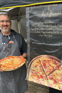 Ein Mann mit Brille steht vor einem Werbeplakat, auf dem eine Pizza abgebildet ist. Er lacht und präsentiert selbst eine Pizza.