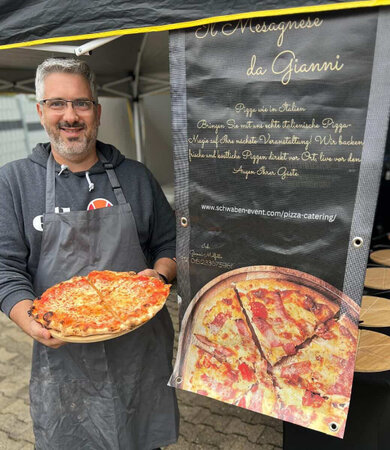 Ein Mann mit Brille steht vor einem Werbeplakat, auf dem eine Pizza abgebildet ist. Er lacht und präsentiert selbst eine Pizza.