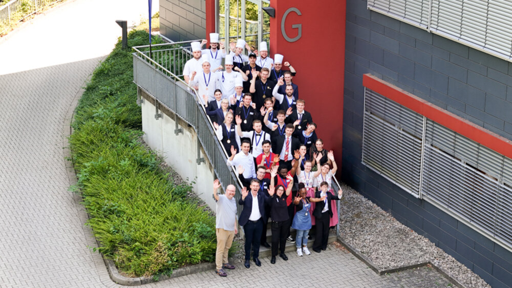 Ein Gruppenfoto auf einer Treppe im Freien vor einem Gebäude: Viele junge Menschen, teils in Berufskleidung des Gastgewerbes, jubeln