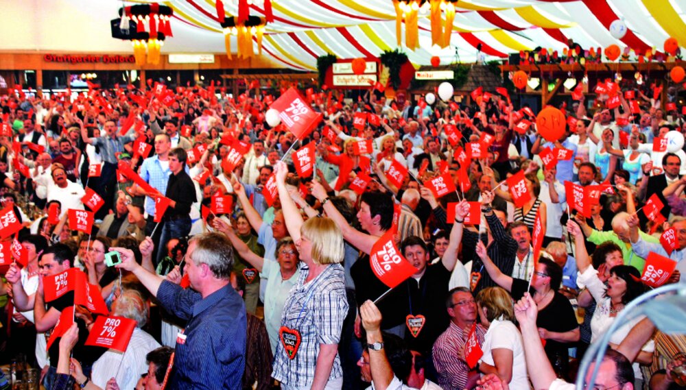 Foto vieler Menschen in einem großen Festzelt, die "Pro 7%"-Fähnchen in den Händen halten.