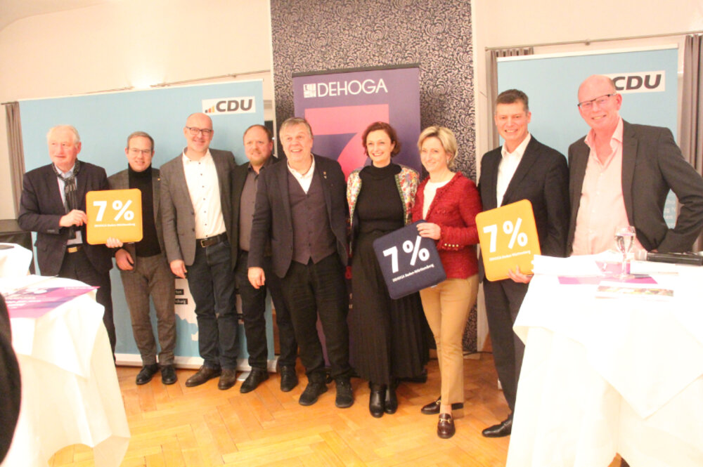Foto von neun Personen, die für die Kamera posieren und Sitzkissen mit der Aufschrift 7% in den Händen halten. Im Hintergrund sieht man DEHOGA- und CDU-Plakate.