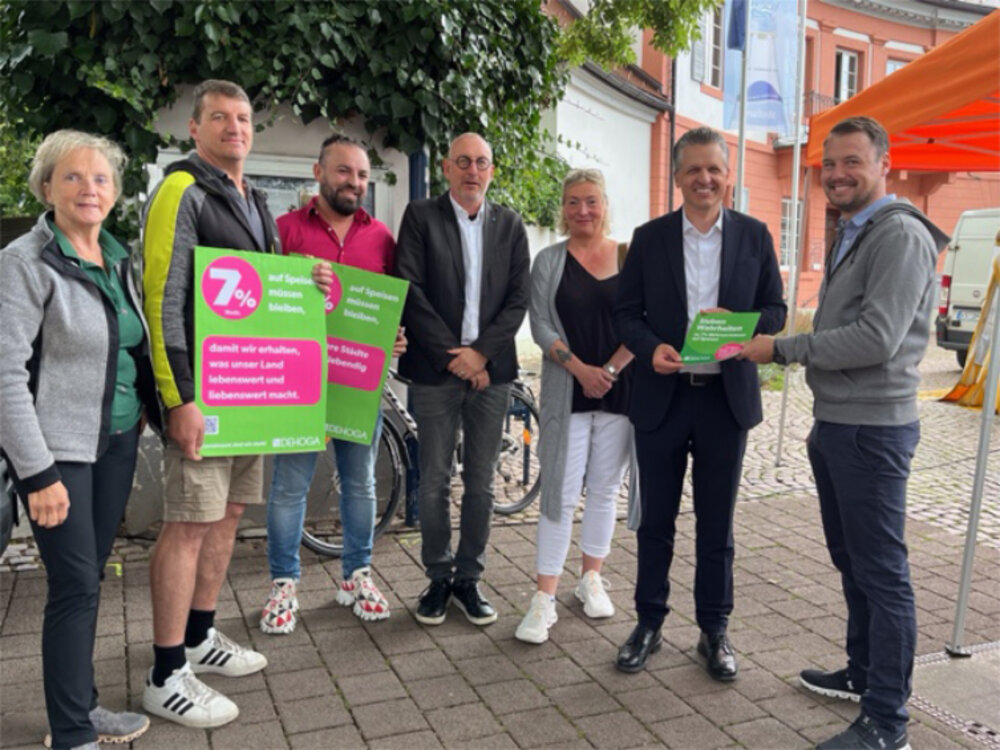 Foto von fünf Männern und zwei Frauen, stehend vor einem Haus. Sie halten 7%-Plakate in den Händen.